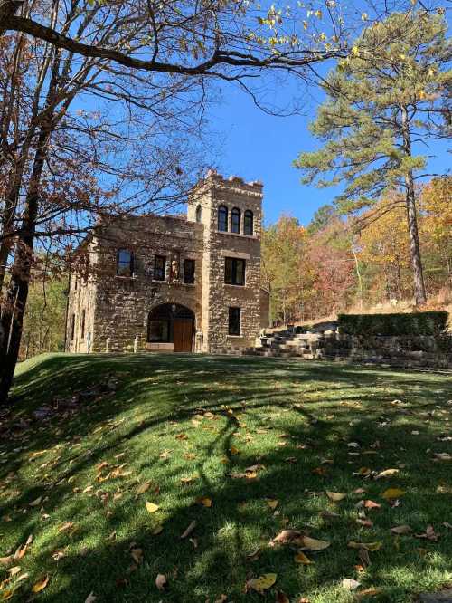 A stone building surrounded by trees and autumn foliage, set on a grassy slope under a clear blue sky.