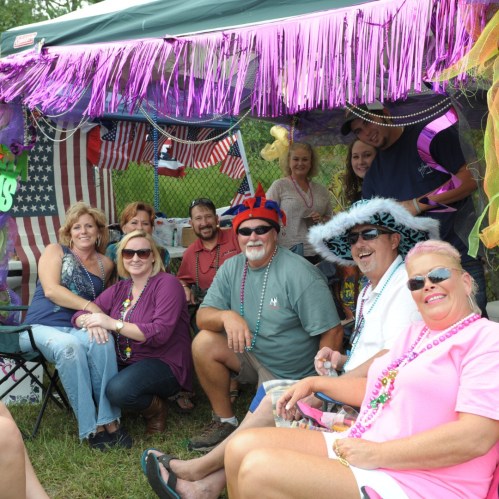 A group of smiling people in festive attire gathered under a decorated tent, enjoying a lively outdoor event.