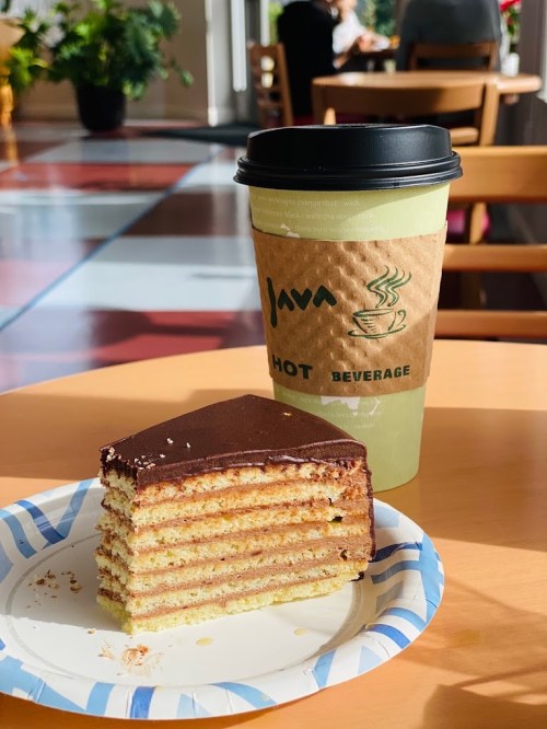 A slice of layered cake on a paper plate next to a coffee cup in a cozy café setting.