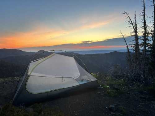 A tent set against a colorful sunset over a mountainous landscape, with trees silhouetted in the foreground.