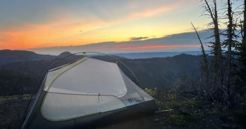 A tent sits on a hillside at sunset, with vibrant colors in the sky and distant mountains in the background.