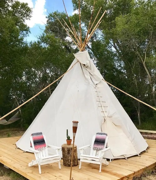 A white teepee on a wooden deck, with two white chairs and a small table, surrounded by trees and blue sky.