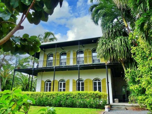 A two-story house with yellow shutters, surrounded by lush greenery and palm trees under a partly cloudy sky.