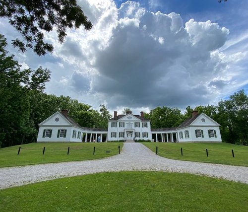 A large white mansion with a gravel driveway, surrounded by green grass and trees under a cloudy sky.