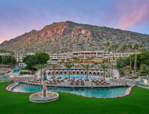 A scenic resort with a pool, palm trees, and mountains in the background under a colorful sky at sunset.