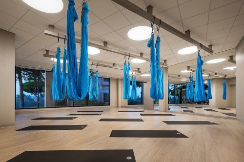 A spacious yoga studio with blue aerial silks hanging from the ceiling and yoga mats arranged on the floor.