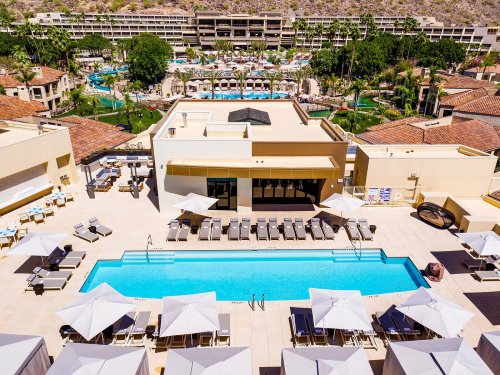 Aerial view of a resort pool surrounded by lounge chairs and umbrellas, with lush landscaping and buildings in the background.