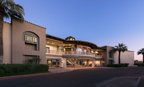 Exterior view of a tavern building at dusk, featuring a balcony and palm trees in the foreground.