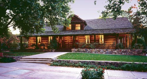 A rustic wooden house with a stone foundation, surrounded by trees and a well-kept lawn, at dusk.