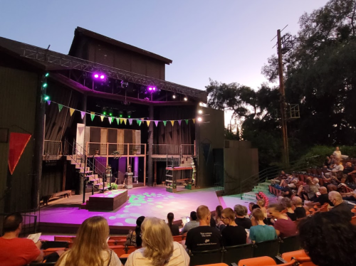 An outdoor theater stage set for a performance, with colorful lights and an audience seated in the foreground.