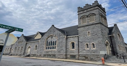 A large stone church building with a tower, featuring arched windows and a cloudy sky in the background.
