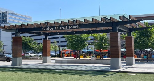 Entrance to Color Burst Park featuring a wooden pergola and green landscaping under a clear blue sky.