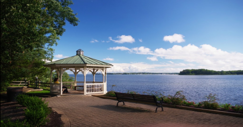 A serene lakeside view featuring a gazebo, bench, and lush greenery under a bright blue sky with scattered clouds.
