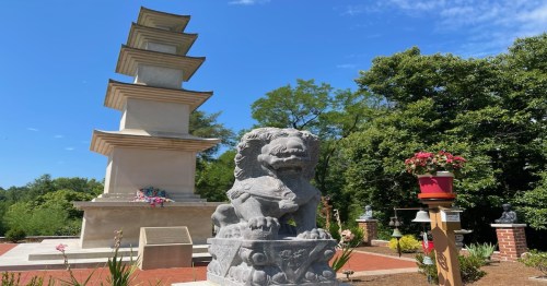 A stone lion statue stands in front of a tall pagoda surrounded by greenery and flowers on a sunny day.
