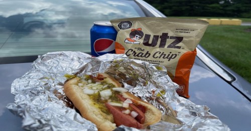 A hot dog with onions and pickles, a bag of Cutz Crab Chips, and a can of Pepsi on a car hood.