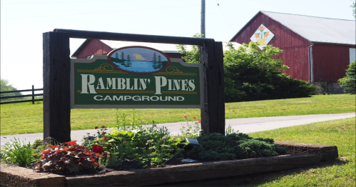 Sign for Ramblin' Pines Campground with flowers in front, set against a backdrop of a red barn and green landscape.