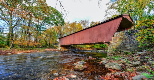 A red covered bridge spans a stream, surrounded by colorful autumn foliage and rocky riverbanks.