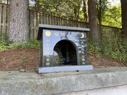 A small stone shrine with inscriptions, surrounded by trees and a wooden fence in the background.