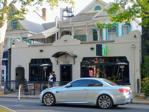 A silver convertible parked in front of a stylish restaurant with outdoor seating and a vintage building.