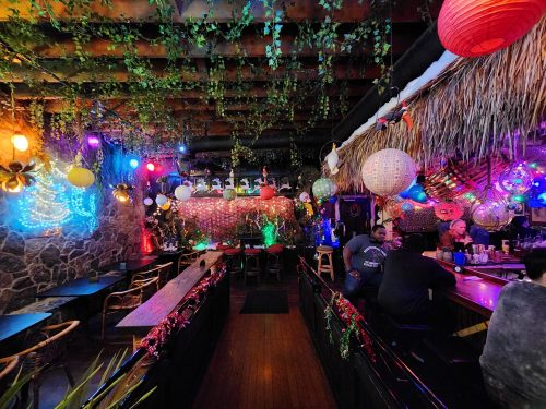 A vibrant bar interior adorned with colorful lights, hanging lanterns, and lush greenery, creating a festive atmosphere.