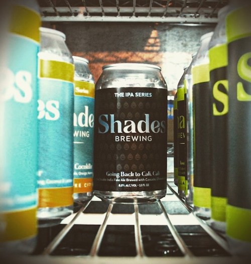 Cans of Shades Brewing beer displayed on a shelf, featuring various colorful labels and a central black can.