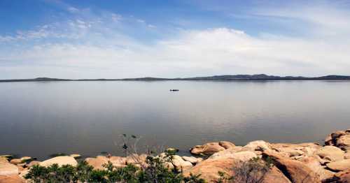 A calm lake with smooth water, rocky shoreline, and distant hills under a clear blue sky. A small boat is visible on the water.