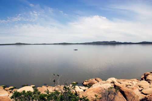 A serene lake scene with smooth water, distant hills, and a small boat in the center, surrounded by rocky shores.