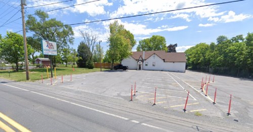 A vacant parking lot next to a white building with a brown roof, surrounded by trees under a clear blue sky.