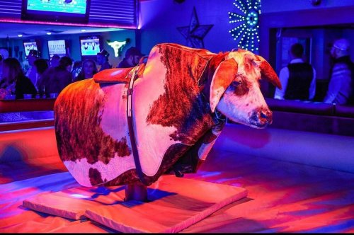A mechanical bull covered in a cowhide blanket, set in a vibrant, colorful bar atmosphere.
