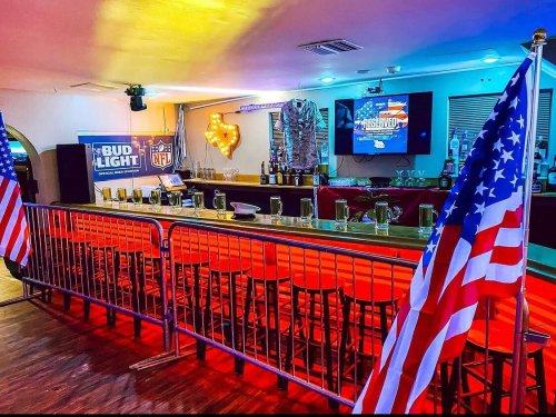 A bar setup with American flags, NFL decor, and a large screen, featuring empty glasses and a vibrant atmosphere.