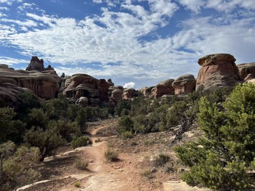 A winding dirt path leads through rocky formations and lush greenery under a cloudy blue sky.