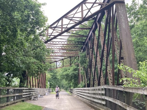 A cyclist rides across a rustic metal bridge surrounded by lush greenery and wooden benches.