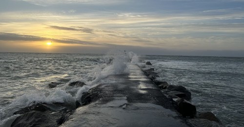 A rocky jetty extends into the ocean at sunset, with waves crashing against it under a colorful sky.