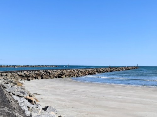 A sandy beach with a rocky jetty extending into calm blue waters under a clear blue sky.