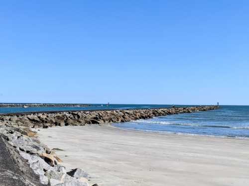 A sandy beach with a rocky jetty extending into calm blue waters under a clear blue sky.