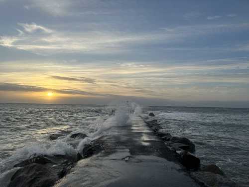 A rocky jetty extends into the ocean at sunset, with waves crashing against it under a colorful sky.