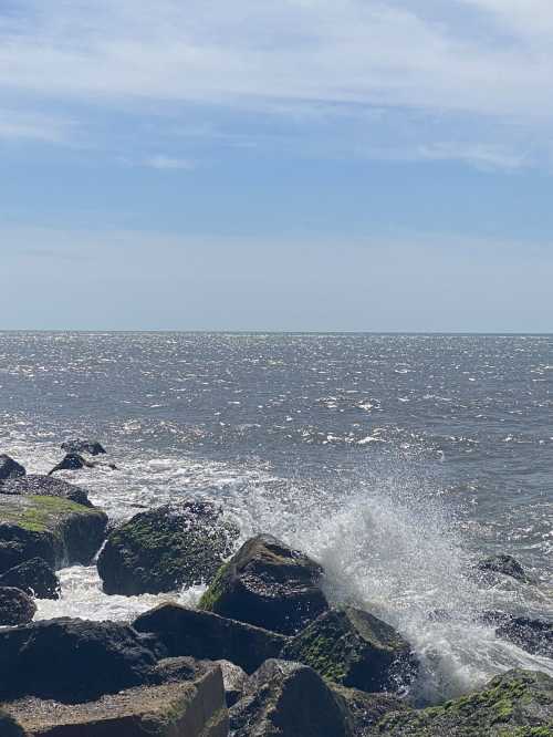 Waves crashing against rocky shore under a clear blue sky with sunlight reflecting on the water.