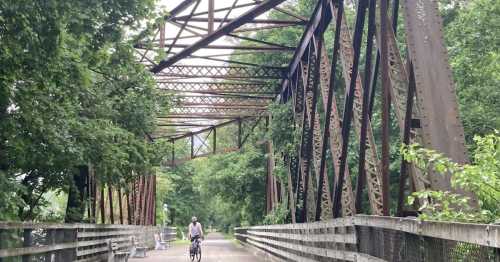 A cyclist rides on a wooden bridge surrounded by lush greenery and a rusted metal structure overhead.