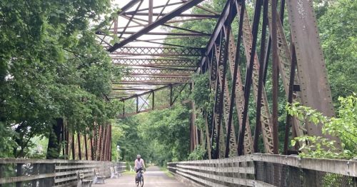 A cyclist rides on a wooden bridge surrounded by lush greenery and a rusted metal structure overhead.