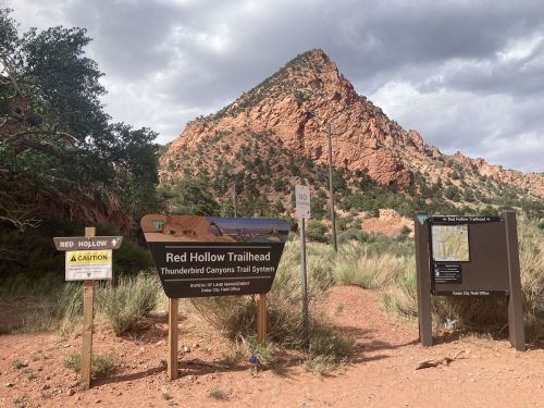 Signage at Red Hollow Trailhead with rocky mountains in the background and desert vegetation. Cloudy sky above.