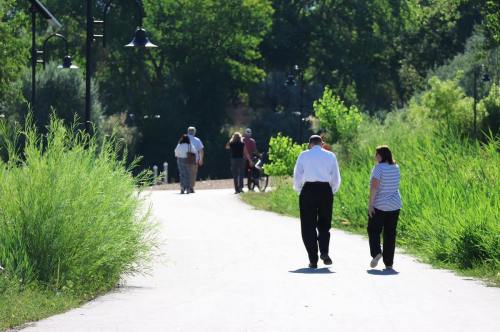 A couple walks on a path surrounded by greenery, with other people in the background enjoying the park.