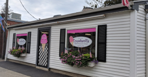 A quaint ice cream shop with pink signage and flower boxes, featuring black shutters and a checkered pattern on the door.