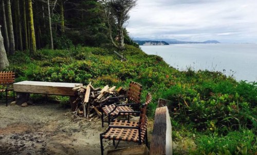 A scenic view of a coastal area with wooden benches and chairs surrounded by greenery and a stack of firewood.