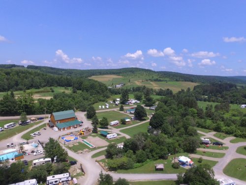 Aerial view of a campground with RVs, a pool, and green hills under a blue sky with fluffy clouds.