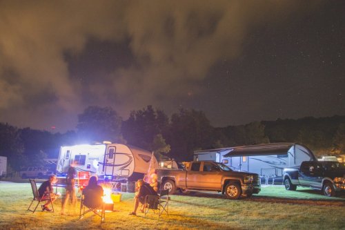 A campsite at night with campers, a fire pit, and vehicles under a starry sky, surrounded by trees.