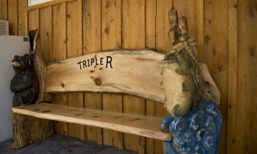A wooden bench with carved bear and fish designs, featuring the name "TRIPLER" on the backrest against a wooden wall.