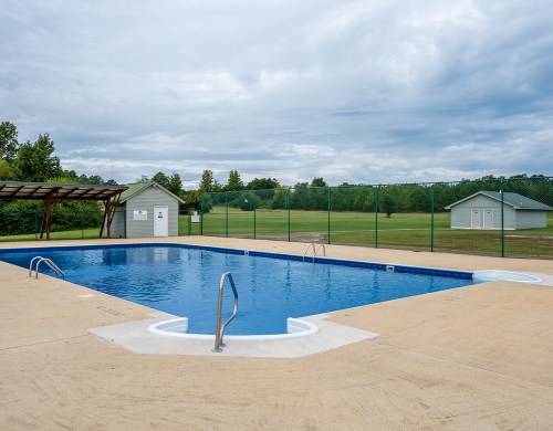 A clear swimming pool surrounded by a concrete deck, with a grassy area and cloudy sky in the background.