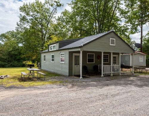 A small green cabin with a porch, surrounded by trees and a gravel path, featuring picnic tables nearby.