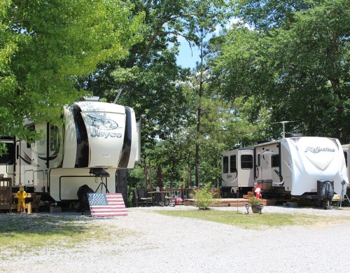 Two RVs parked in a shaded area, surrounded by trees and gravel, with an American flag displayed nearby.