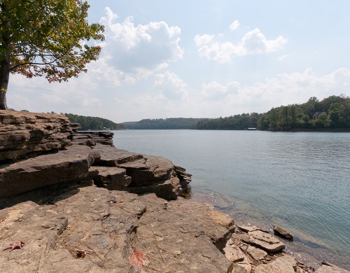 A serene lakeside view with rocky shorelines, trees, and a cloudy sky reflecting over calm water.